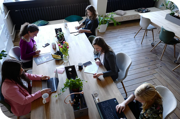 Professional team collaborating at a large shared workspace An overhead, high-angle view of five women working around a long wooden conference table in a modern, sunlit co-working space. Several team members are using laptops, while others review printed documents and take notes.