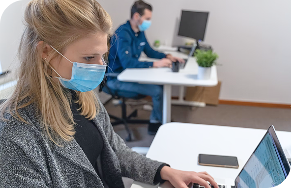 Office employees working at desks while wearing protective masks. A close-up side view of a blonde woman wearing a blue surgical face mask while typing on a laptop at her office desk. In the blurred background, a male colleague also wearing a mask works at a separate workstation.
